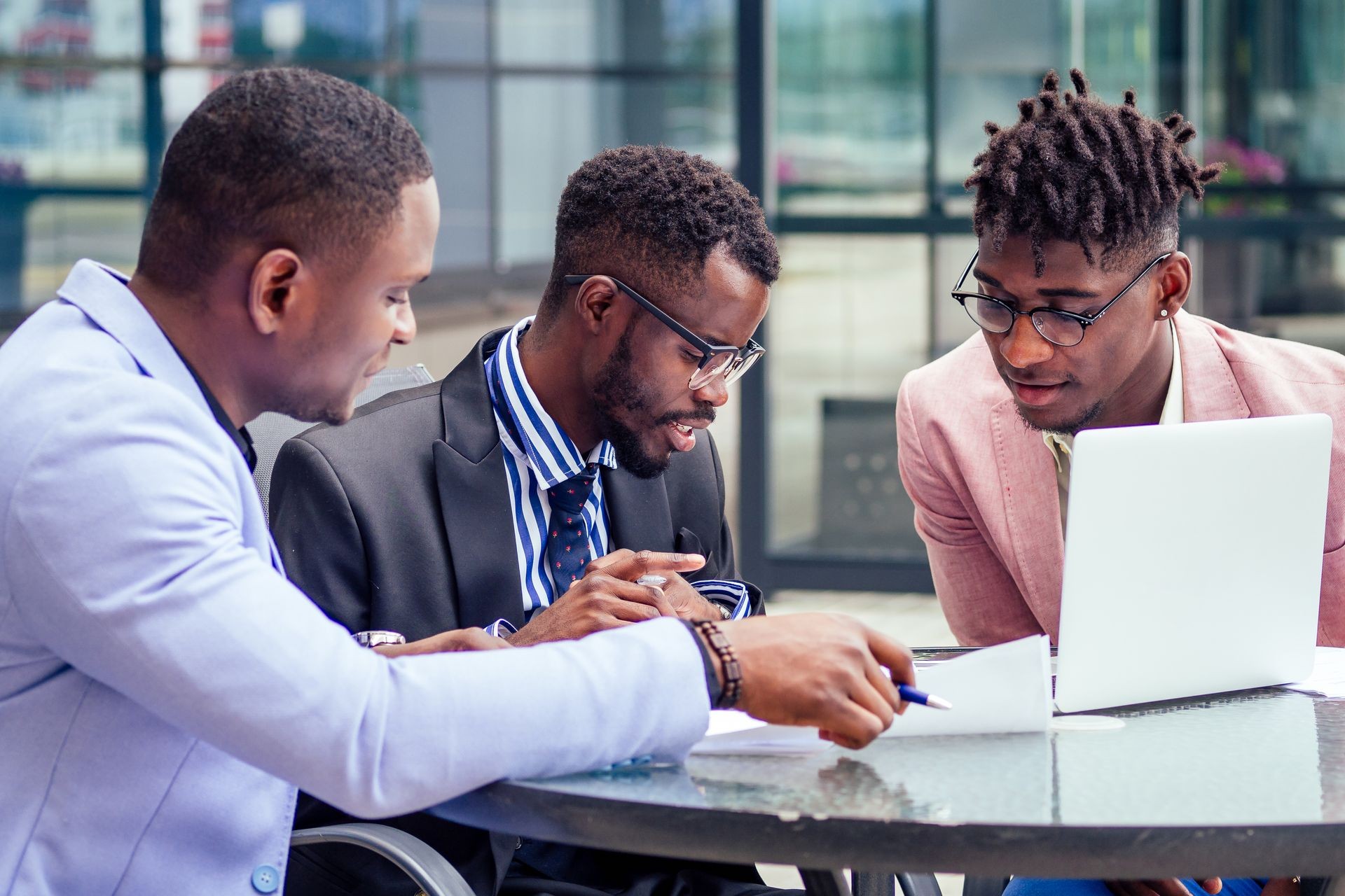 A group of three stylish African American businessman partners entrepreneurs in fashion business suits meeting sitting at table sign securities with laptop in a summer cafe outdoors